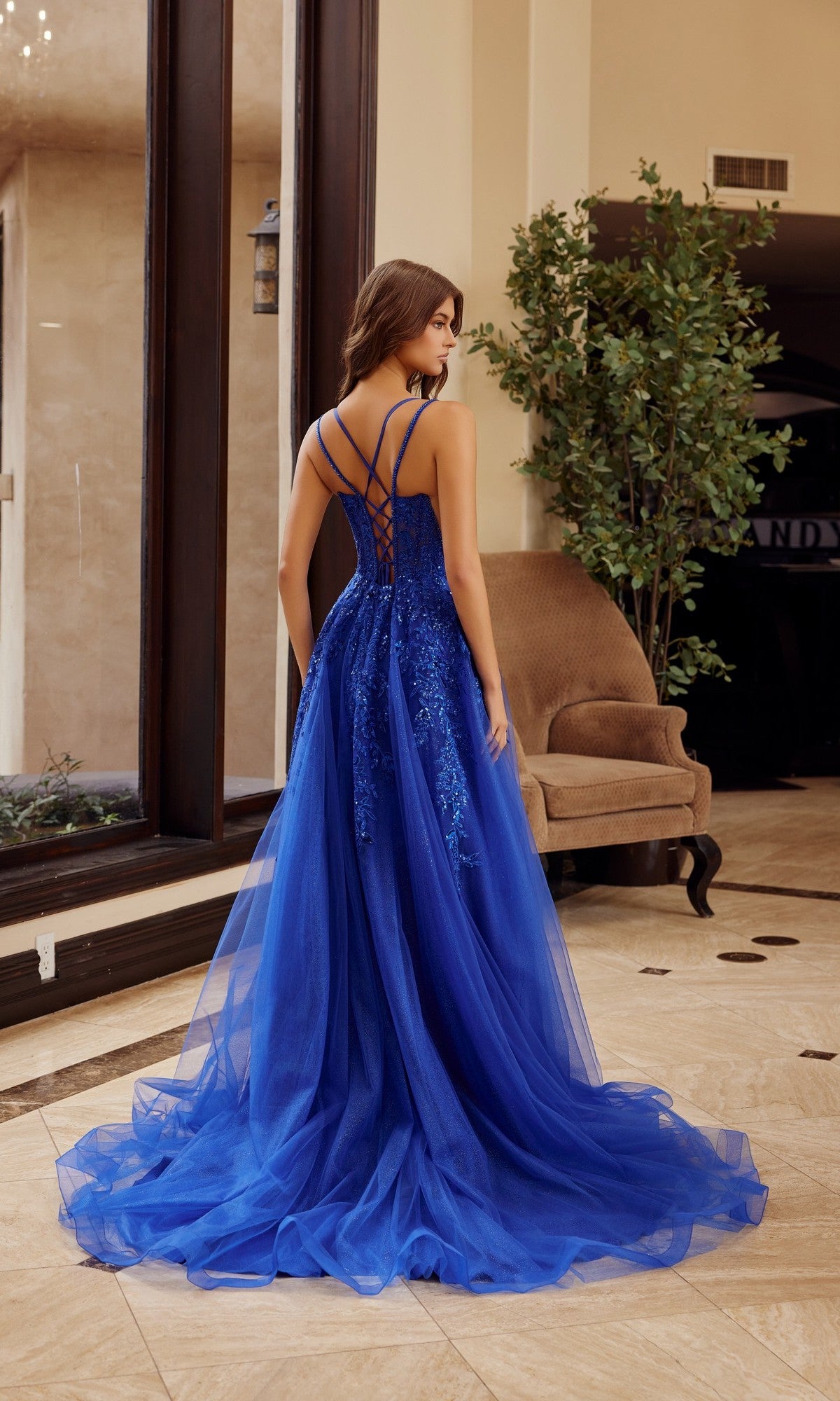 A woman stands indoors wearing the Nox Anabel H1885 long prom dress in royal blue, featuring thin straps, a corset bodice, open back, and lace-tulle details.